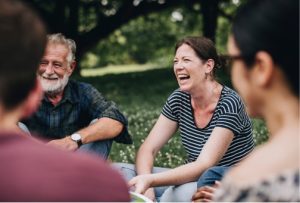 A candid moment of an older man with a beard and a smiling woman in a striped top enjoying a conversation with others in a park setting.